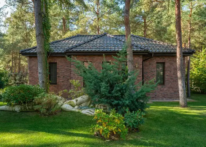 Single-storey brick bungalow with a hipped tiled roof set among tall pine trees and a lawn