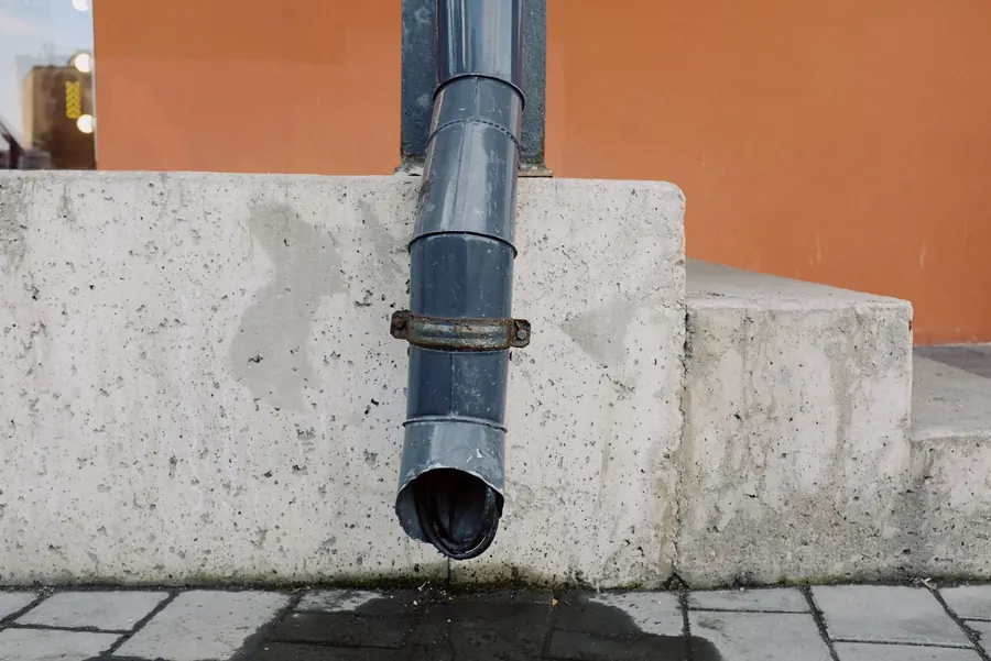 Rainwater dripping from the end of a downpipe beside concrete steps, showing where a flat roof outlet discharges