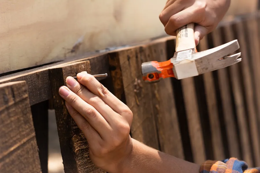 Hands driving a nail into a dark timber board with an orange-handled claw hammer