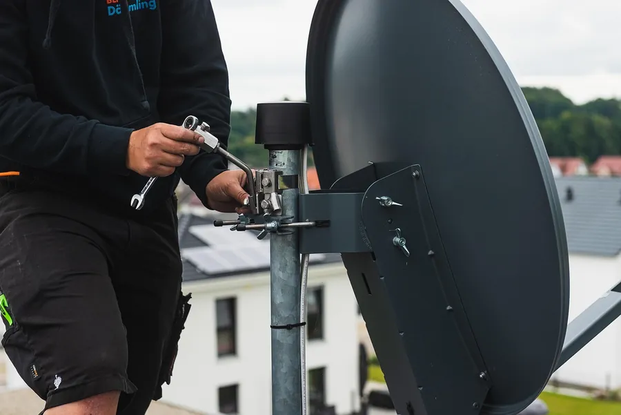 Installer tightening the bracket of a grey satellite dish on a pole, with houses in the background