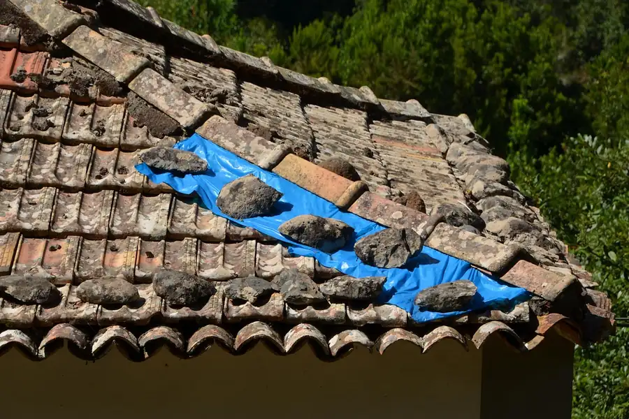 Damaged clay tile roof with broken tiles and a blue tarpaulin weighed down with stones as a stopgap repair