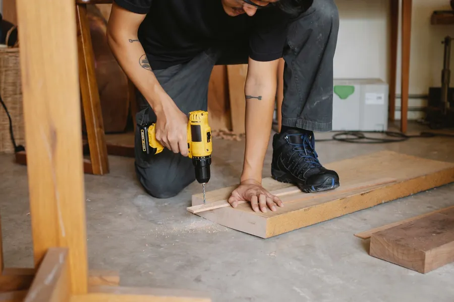 Tradesman kneeling to drive a screw through a timber board with a cordless drill
