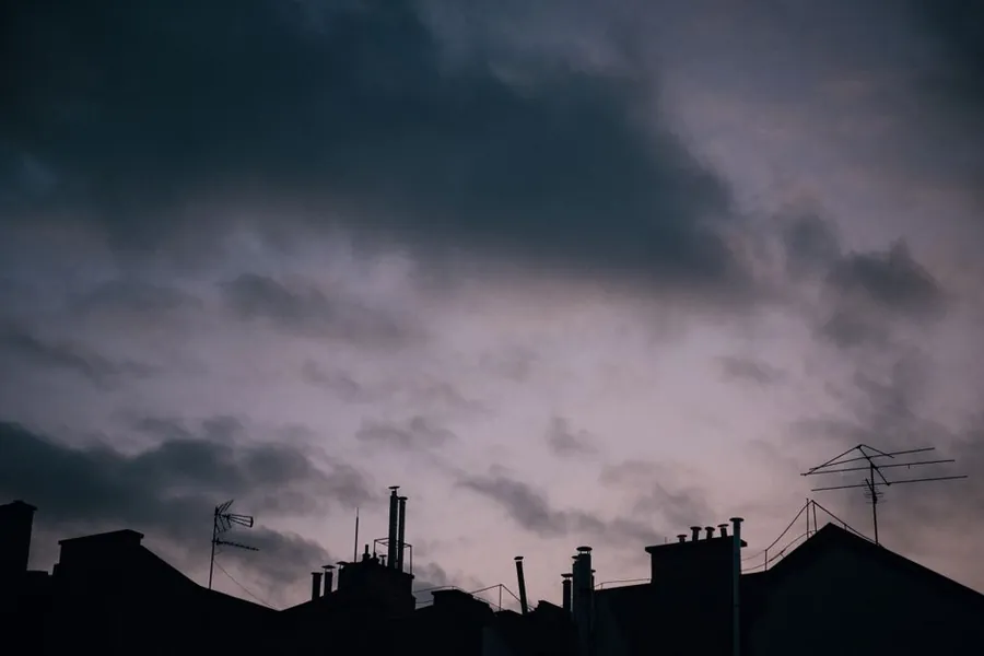 Silhouetted rooftops chimneys and television aerials under a dark storm sky