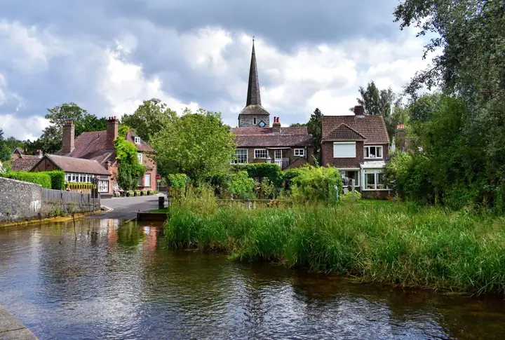 Village houses and a church spire beside a slow river, similar to the old core of Ollerton by the River Maun