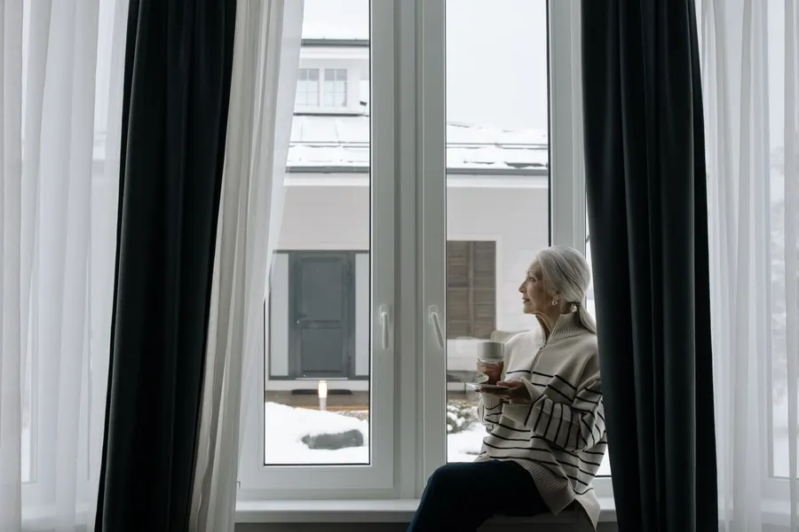 Older woman sitting by a window with a warm drink in a cold home during winter