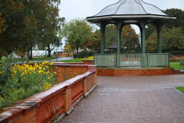 A green-painted bandstand behind a red brick balustrade and flower beds in an English town park, much like Kingsway Park in Kirkby-in-Ashfield