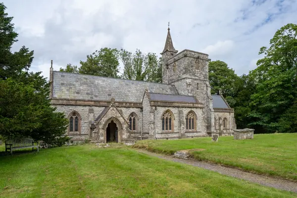 Grey stone village church with a slender spire and slate roofs set in a grassy churchyard under a cloudy sky