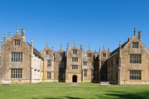 Honey-coloured stone country house with tall gables and mullioned windows behind a level lawn, typical of a ducal estate