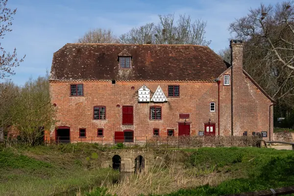 Red brick watermill with a steep clay tiled roof beside a river in the English countryside, similar to the listed mill in Old Ollerton
