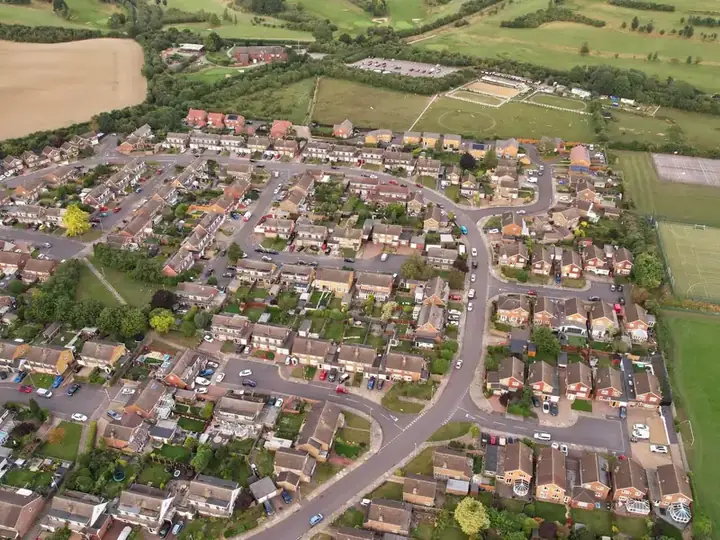 Aerial view of a brick-built English village estate with tiled roofs, surrounded by fields and woodland, like Rainworth on the edge of Sherwood
