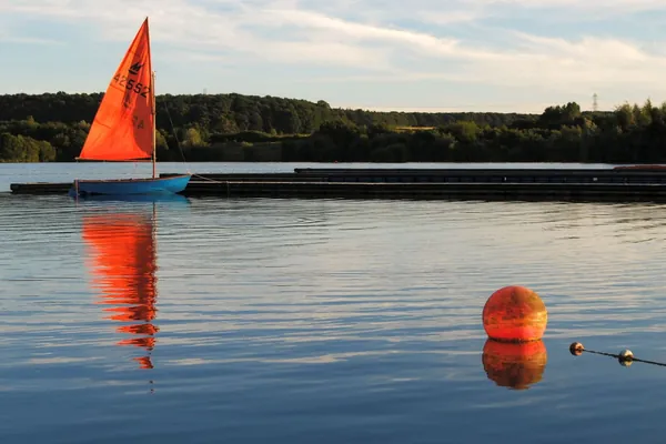 A small dinghy with an orange sail moored on calm water at evening, a sailing scene like those on Kings Mill Reservoir near Sutton-in-Ashfield